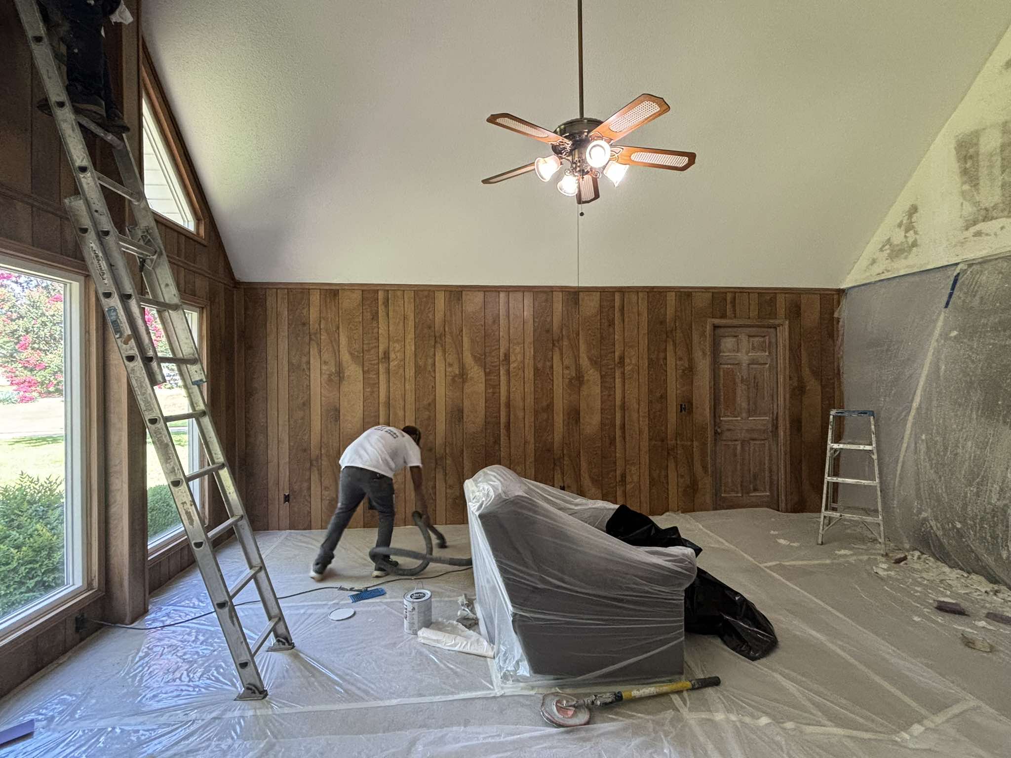 Vaulted room during preparation with wood-paneled walls, protected floors, ladders, and a painter at work